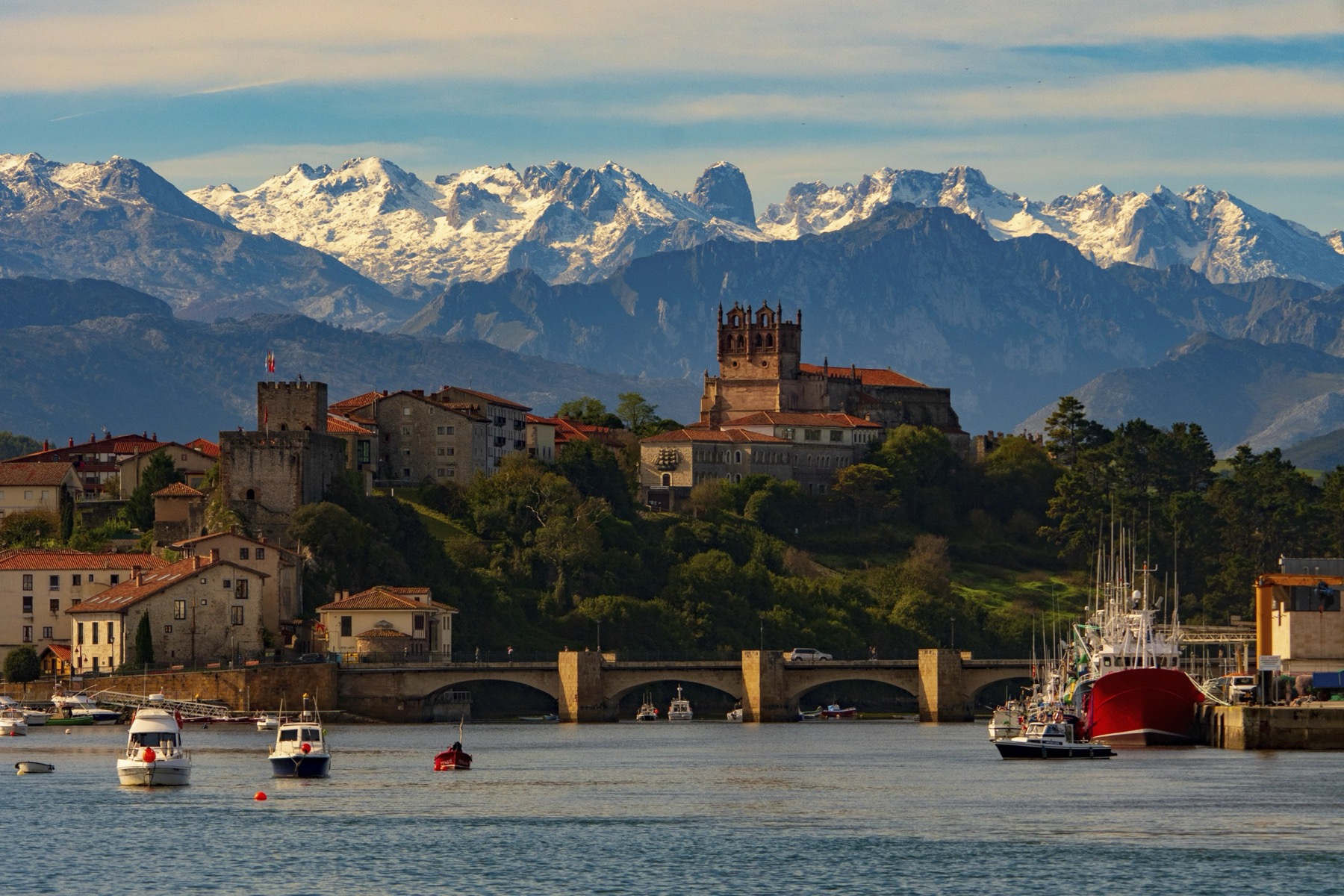 San Vicente de la Barquera harbour at golden hour, with the snow-capped peaks of the Picos de Europa rising behind the medieval town and its cathedral.