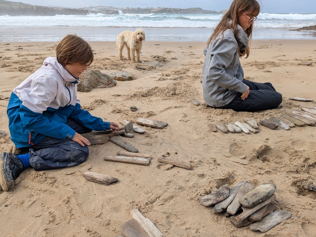 Rafa and Alba kneeling on a wide Cantabrian beach at low tide, sorting through piles of weathered driftwood pieces — keyfob candidates. Loki the Labradoodle stands in the surf behind them, watching.