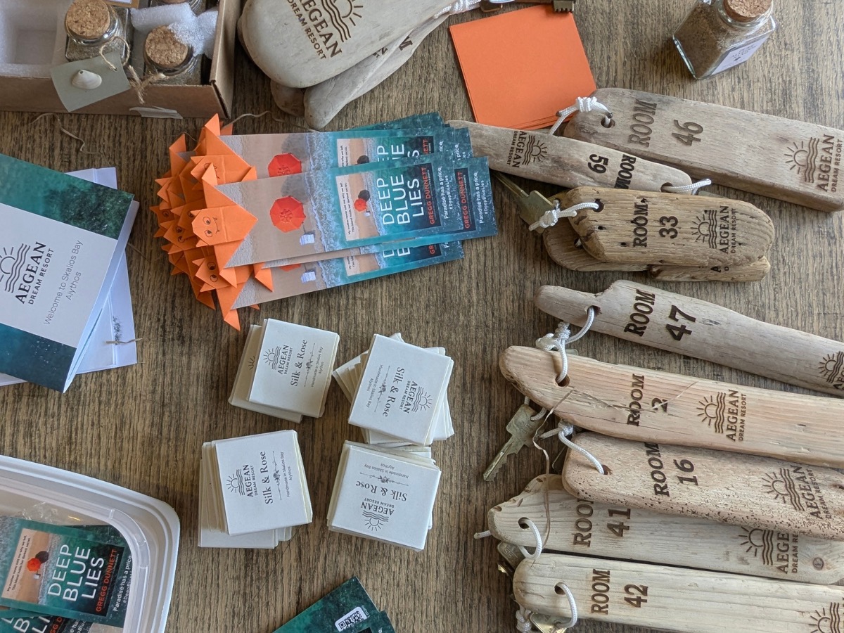 An overhead flat-lay of the box contents in production: small glass jars of Alythos sand with shell tags, a stack of orange origami bookmarks topped with Deep Blue Lies cards, stacks of 'Silk & Rose' Aegean Dream Resort soap, and a row of laser-engraved driftwood room keyfobs — Rooms 46, 59, 33, 47, 16, 41, 42.
