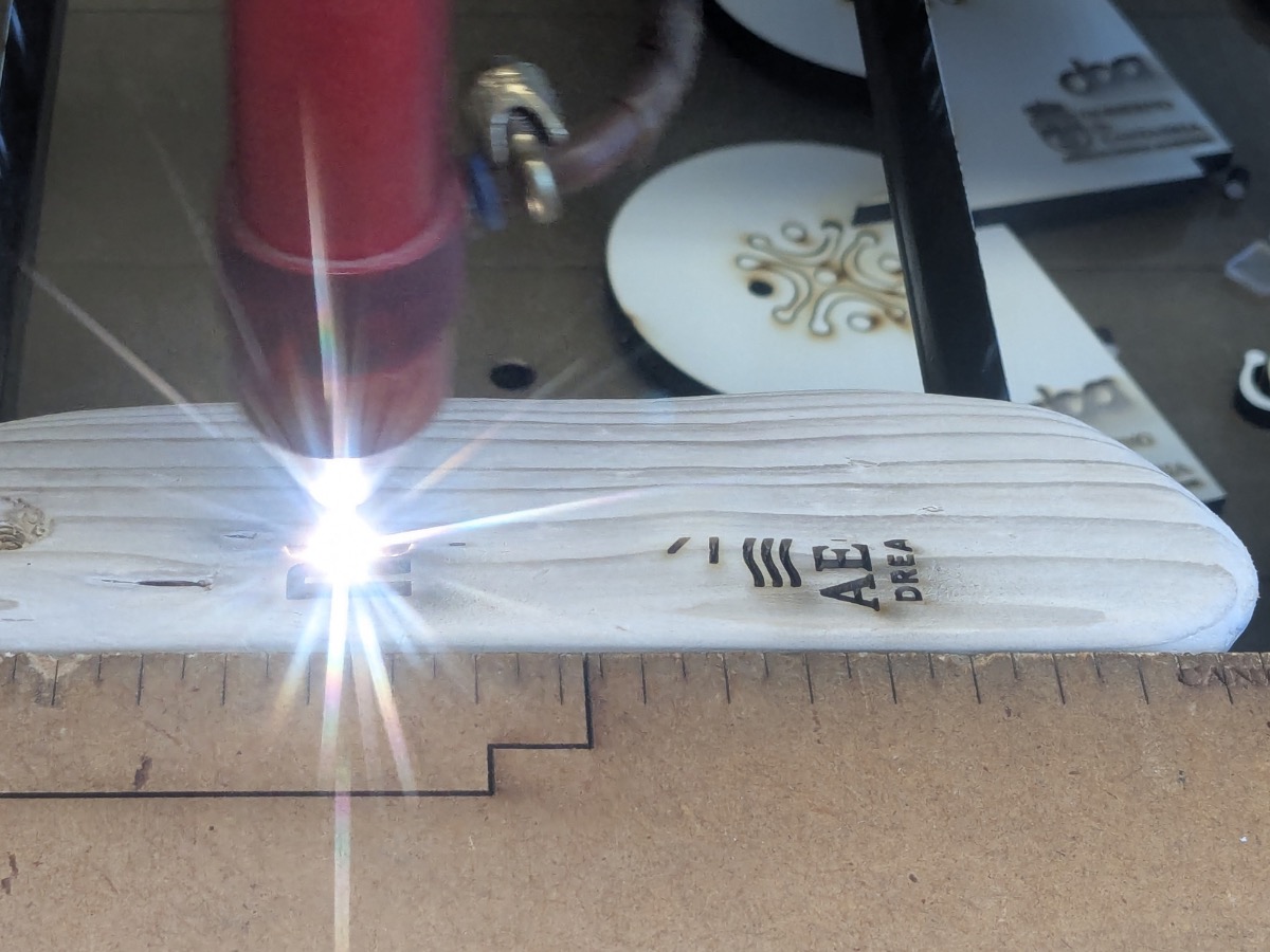 Close-up of a CO2 laser engraver mid-burn on a sanded driftwood keyfob — bright spark from the laser tip, partial AE letters of the Aegean Dream Resort logo emerging in the wood, a finished filigree-engraved piece visible in the background.