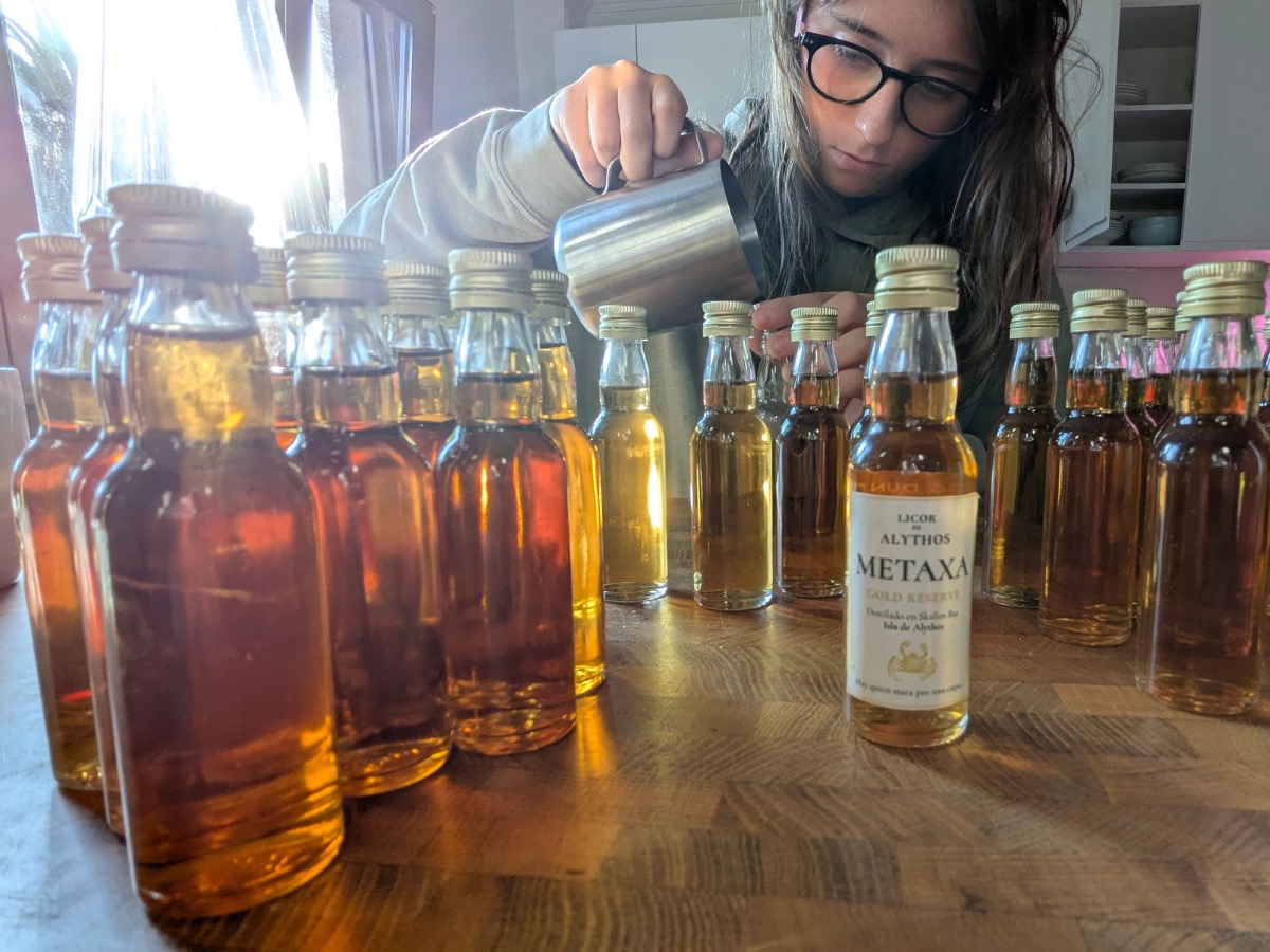 Alba, head down in concentration, decanting Metaxa from a hip-flask sized vessel into a row of miniature spirit bottles lined up on the kitchen table.