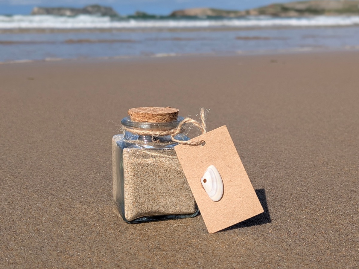 A small square glass jar of Alythos sand with a cork stopper, sitting on a wet Cantabrian beach. Tied to its neck by twine, a kraft-card tag with a single white shell pinned to it — the shell has a neat round hole drilled clean through by an octopus.