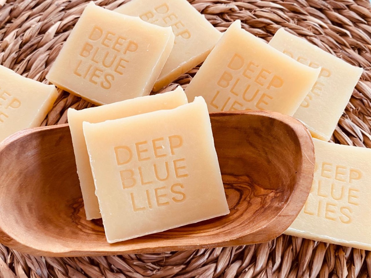 A pile of hand-made artisan soap blocks stamped with 'DEEP BLUE LIES', resting in and around an olive-wood dish on a woven reed mat.
