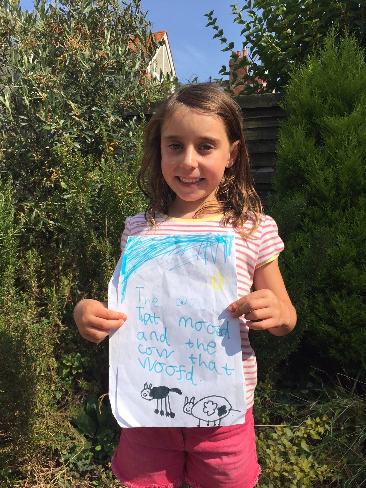 Alba, age six, in the garden, holding up the hand-drawn cover for her shopkeeper's book The Dog that Mooed and the Cow that Woofd.